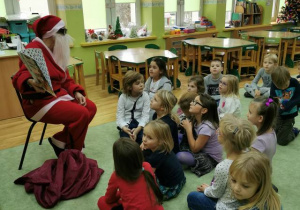 Dzieci siedzą na dywanie, na krześle siedzi Mikołaj, pokazuje dzieciom obrazki w książce